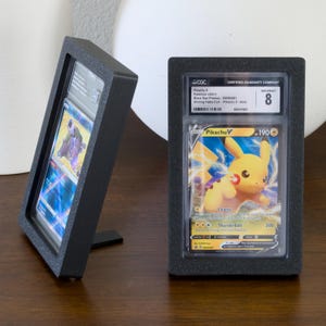 May include: Two black display cases holding graded Pokémon cards. One case is angled, showcasing a card with a blue and purple design. The other case displays a Pikachu V card, graded NM/MINT 8 by CGC, with the text "Charge" and "Thunderbolt".