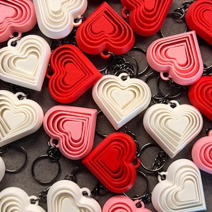 May include: A collection of layered heart-shaped keychains in red, pink, and white. The keychains are made of plastic and have a layered design, with each layer slightly smaller than the one before it. The keychains are a fun and festive way to show your love for Valentine's Day.