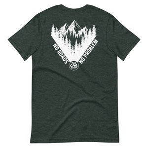 以下が含まれることがあります： 暗い緑色のTシャツに、白い山脈と木々のグラフィックが描かれており、三角形の中に「No Roads No Problem」と「The Car Show」の文字が書かれています。