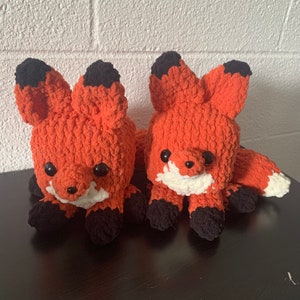 May include: Two crocheted fox plushies with orange and white fur. The foxes have black eyes and noses and are sitting on a dark surface.
