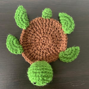 Puede incluir: Un juguete de tortuga de crochet con una concha marrón y extremidades verdes. La tortuga tiene una cabeza pequeña y redonda con ojos negros.
