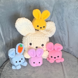 Puede incluir: Cuatro peluches de conejo de crochet en diferentes colores. Un conejo blanco con un conejo amarillo en la parte superior, un conejo azul claro con una zanahoria, un conejo morado claro y un conejo rosa.