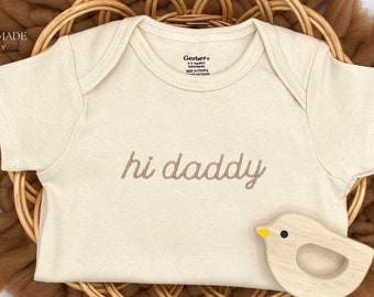 May include: A cream-colored baby onesie with the text "hi daddy" embroidered in brown on the front. The onesie is lying on a brown woven basket with a wooden bird toy.