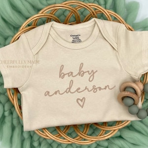 May include: A beige baby onesie with the text "baby anderson" embroidered in brown cursive script with a heart underneath. The onesie is on a woven wicker basket with a green fuzzy blanket.
