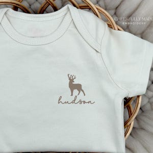 May include: Cream-colored baby onesie with a brown deer graphic and the name "hudson" in a script font. The onesie is short-sleeved and has a round neckline. The words "CHEERFULLY MADE EMBROIDERY" are in the upper right corner.