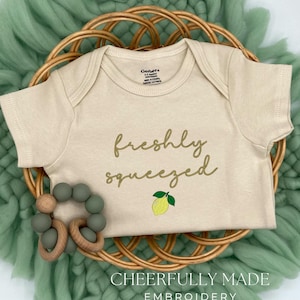 May include: A beige baby onesie with the words "freshly squeezed" embroidered in gold script, along with a lemon graphic. A wooden and green bead teether is next to the onesie, all on a woven basket and green fabric. The text "CHEERFULLY MADE EMBROIDERY" is at the bottom.