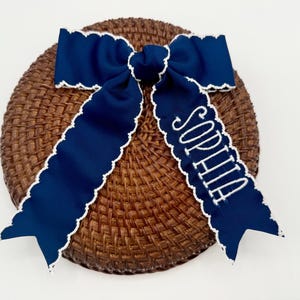 May include: A navy blue ribbon bow with white scalloped edges, resting on a woven brown surface. The bow features the name "SOPHIA" embroidered in white. A decorative hair accessory.