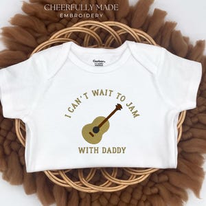 May include: White baby onesie with the embroidered text "I CAN'T WAIT TO JAM WITH DADDY" and a guitar graphic. The onesie is displayed on a woven, circular, brown background.