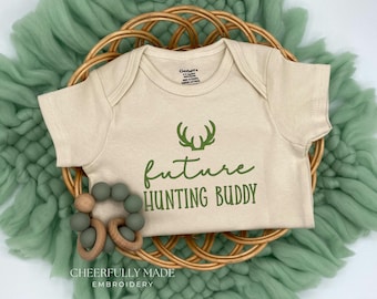 May include: A cream-colored baby onesie with green text that reads "future hunting buddy" and a green silhouette of antlers above the text.