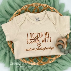 May include: A beige baby onesie with the text "I rocked my session with (custom photography)" in brown lettering. The onesie is on a woven basket with a green, textured background. A wooden teething ring and a beaded teether are on the right.