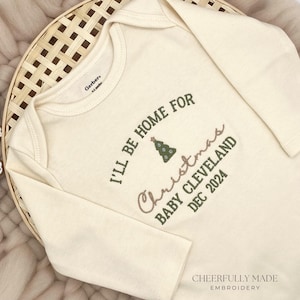 May include: A cream-coloured long-sleeved onesie with the text "I'll be home for Christmas Baby Cleveland Dec 2024" embroidered in green and brown. The onesie has a small green Christmas tree design.