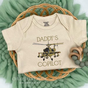 May include: A beige baby onesie with the text "DADDY'S COPILOT" and a helicopter graphic. The onesie is displayed on a woven basket, with a green wool background and a wooden teething ring.