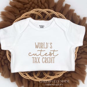 May include: White baby onesie with the text "World's Cutest Tax Credit" in a brown script font. The onesie is displayed on a brown woven circular base with a white background. The brand name "Gerber" is visible.