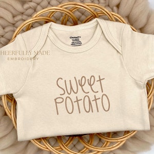 May include: A beige baby onesie with the text "Sweet Potato" embroidered in brown on the front. The onesie is lying on a woven basket.