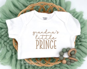 May include: White baby onesie with the text "grandma's little PRINCE" in brown script. The onesie is displayed on a woven basket with a green textile background. A wooden and green bead teether is next to the onesie.