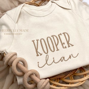 May include: A cream-colored baby onesie with the text "Cheerfully Made Embroidery" and the names "Kooper Ilian" embroidered in brown thread.
