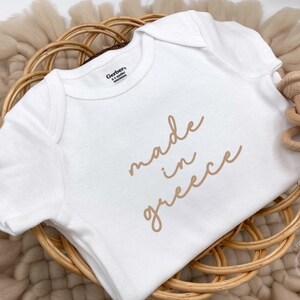 May include: A white baby onesie with the words "made in greece" in a light brown script. The onesie is on a woven basket with a wooden rattle. The onesie is a Gerber brand, size 0-3 months.