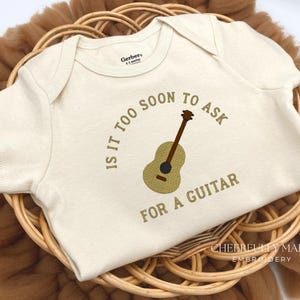 May include: Cream-colored baby onesie with a guitar graphic and the text "IS IT TOO SOON TO ASK FOR A GUITAR". The onesie is displayed in a woven basket.