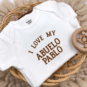 May include: White baby onesie with the text "I LOVE MY ABUELO PABLO" in brown lettering. A wooden baby rattle is next to the onesie, which is resting on a woven basket. The onesie is a Gerber brand, 0-3 months.