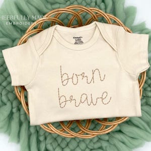 May include: A cream-colored baby onesie with the embroidered words "born brave" in a cursive font. The onesie is displayed on a woven, circular basket, set against a soft, green, textured background. The text "Cheerfully Made Embroidery" is in the upper left corner.