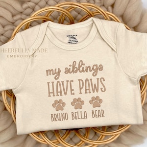 May include: A beige baby onesie with the text "my siblings have paws" and the names "Bruno Bella Bear" underneath. Three paw prints are between the text and the names.