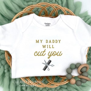 May include: White baby onesie with the text "MY DADDY WILL cut you" in gold lettering, along with a comb and scissors graphic. The onesie is displayed on a woven basket with a green textile background and a wooden and silicone teething ring.