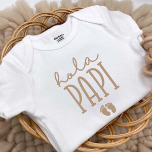 May include: White baby onesie with the words "Hola Papi" in a tan color, along with two small footprints. The onesie is placed in a woven basket with a wooden rattle on the side. The onesie is a Gerber brand.