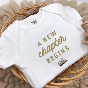 May include: White baby onesie with the text "A New Chapter Begins" in gold script. The onesie is placed in a woven basket with a wooden rattle. The onesie has a small illustration of an open book below the text.