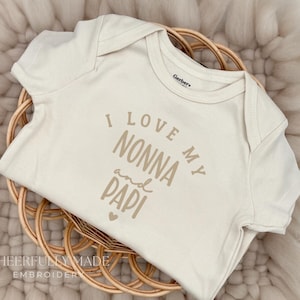 May include: A cream-colored baby onesie with the text "I love my Nonna and Papi" in a brown font. The onesie is folded and placed in a woven basket, set against a soft, neutral-toned background.