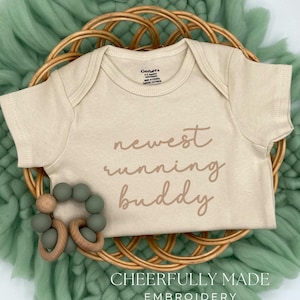 May include: A beige baby onesie with the words "newest running buddy" in a brown script. The onesie is on a woven basket with a green teething ring and a green, woven background. The text "CHEERFULLY MADE EMBROIDERY" is at the bottom.
