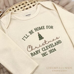 May include: A cream-coloured long-sleeved onesie with the text "I'll be home for Christmas Baby Cleveland Dec 2024" embroidered in green and pink. A small green embroidered Christmas tree is above the text.
