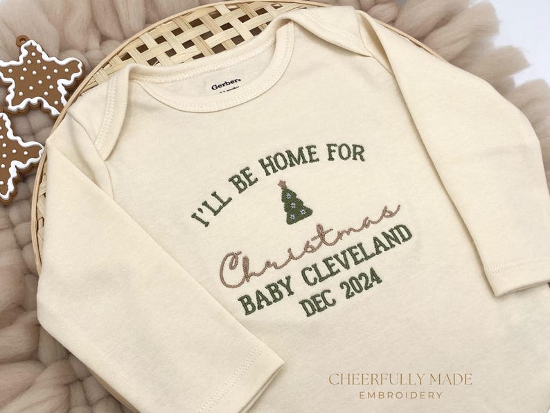 May include: A cream-coloured long-sleeved onesie with the text "I'll be home for Christmas Baby Cleveland Dec 2024" embroidered in green and pink. A small green embroidered Christmas tree is above the text.