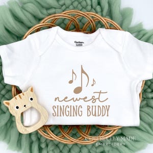 May include: White baby onesie with brown musical notes and the text "newest SINGING BUDDY." A wooden cat-shaped teether sits next to the onesie, which is on a woven basket and green fabric.