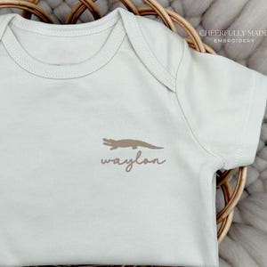 May include: Cream-colored baby onesie with a brown alligator graphic and the name "Waylon" in a matching script font. The onesie is displayed on a woven basket.