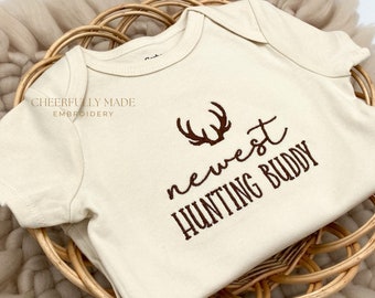 May include: A cream-colored baby onesie with brown embroidery that reads "newest hunting buddy" with a pair of antlers above the text.