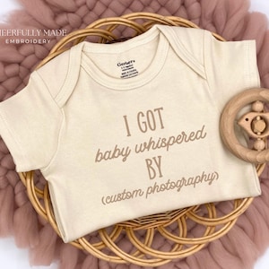 May include: A cream-colored baby onesie with the text "I GOT baby whispered BY (custom photography)". The onesie is on a woven basket with a wooden bird rattle. The background is a pink, textured fabric.