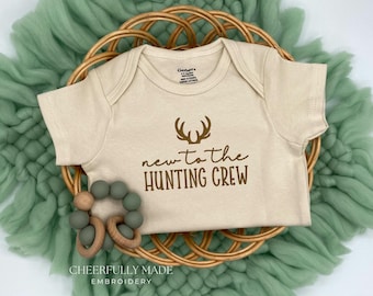 May include: A beige baby onesie with the text "new to the HUNTING CREW" and a deer antler graphic. A wooden and green bead teether is next to the onesie, which is on a woven basket. The background is a soft, green fabric.