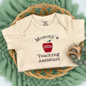 May include: A white baby onesie with the text "Mommy's little Teaching Assistant" embroidered on it. A red apple with a green stem is part of the design. The onesie is lying on a woven basket with a green fuzzy blanket.