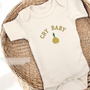 May include: Cream-colored baby onesie with the embroidered text "CRY BABY" above a yellow onion illustration. The onesie is displayed in a woven basket. The text and onion are in shades of yellow and green.
