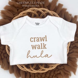 May include: White baby onesie with the words "crawl walk hula" in a brown script. The onesie is centered on a woven, circular, brown decorative piece. The words are in a playful font.
