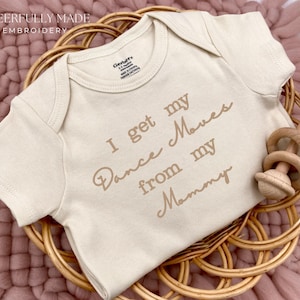 May include: A beige baby onesie with the text "I get my Dance Moves from my Mommy" in brown script. The onesie is in a woven basket with a wooden rattle. The words "CHEERFULLY MADE EMBROIDERY" are at the top.