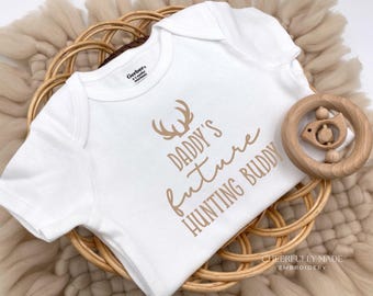 May include: White baby onesie with the text "DADDY'S future HUNTING BUDDY" in brown script and antlers graphic. A wooden baby rattle is next to the onesie. The onesie is on a woven basket.