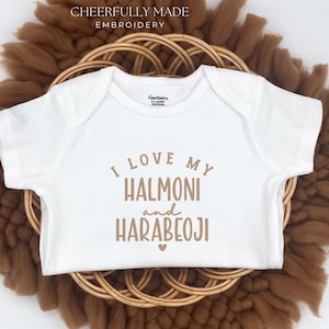 May include: White baby onesie with the text "I LOVE MY HALMONI and HARABEOJI" in brown lettering. The onesie is placed on a woven basket and a brown, fluffy surface. The words "CHEERFULLY MADE EMBROIDERY" are at the top.