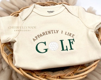 Pode incluir: Um macacão de bebê de cor creme com o texto "Cheerfully Made Embroidery" e "Apparently I Like Golf" bordado em fio marrom e verde. Uma bola de golfe branca é bordada no centro do texto "Golf".