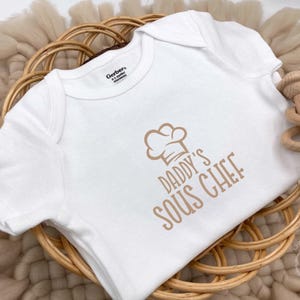 May include: White baby onesie with the text "Daddy's Sous Chef" and a chef hat graphic in a light brown color. The onesie is placed in a woven basket with a wooden rattle.