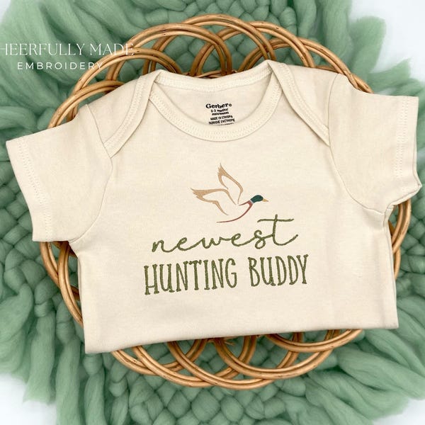 May include: A cream-coloured baby onesie with short sleeves features a brown duck flying with a green head and the text "newest hunting buddy" in brown.