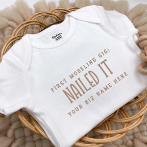 May include: White baby onesie with the text "FIRST MODELING GIG: NAILED IT YOUR BIZ NAME HERE" in brown lettering. The onesie is on a woven basket with a wooden rattle. The onesie is Gerber brand.