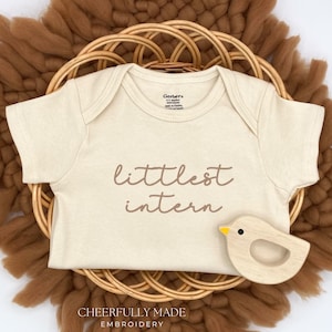 May include: A beige baby onesie with the words "littlest intern" in brown script is centered on a woven basket. A wooden bird toy with a yellow beak rests to the right. The background features a brown, textured, circular mat.