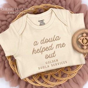 May include: A beige baby onesie with the text "a doula helped me out" and "Golden Doula Services" in brown lettering. A wooden bird-shaped rattle sits beside the onesie on a woven basket, atop a pink textile.