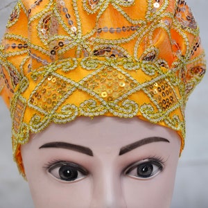 Puede incluir: Un tocado naranja con intrincados detalles de lentejuelas y bordados dorados y plateados. El tocado se exhibe en una cabeza de maniquí, mostrando el diseño y la textura de la tela.